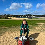 child, playground, spring_rocker, sand, grass, blue_sky, clouds, happy, curly_hair, casual_clothing, outdoor, daylight, park, play_equipment, water, bench, trees, residential_area, shadow, fun