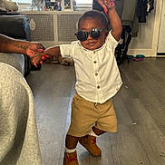 Dax joined the competition — help win amazing prizes! toddler, child, sunglasses, standing, fashion, portrait, smiling, indoor, living_room, adult_hands, white_shirt, brown_shorts, boots, wood_floor, furniture, couch, radiator, window, clothing, family