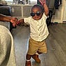 toddler, child, sunglasses, standing, fashion, portrait, smiling, indoor, living_room, adult_hands, white_shirt, brown_shorts, boots, wood_floor, furniture, couch, radiator, window, clothing, family