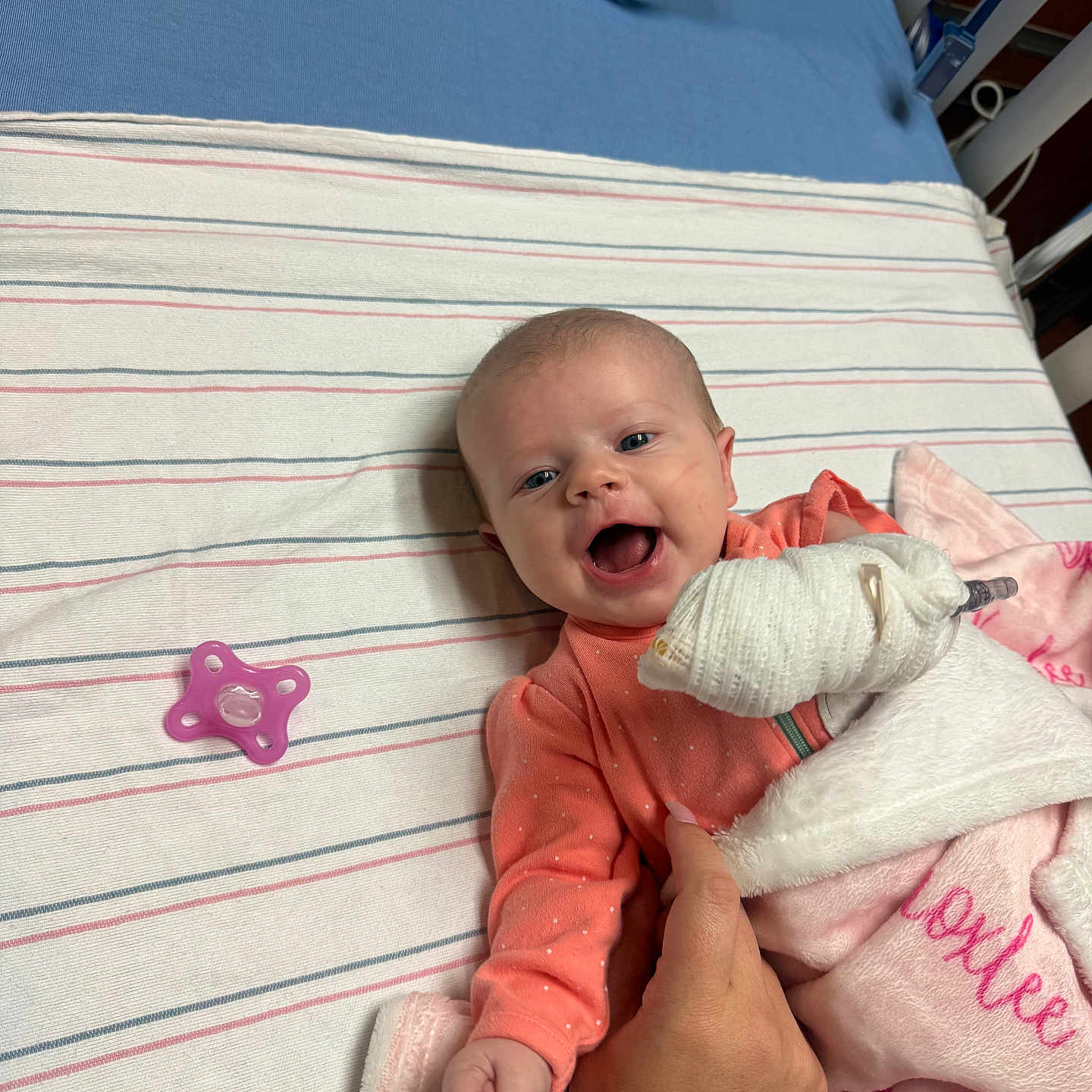 Knoxlee is registered to the contest to win money with this photo: baby, infant, smiling, hospital, bandage, blanket, pacifier, hand, bed, striped_sheet, person, child, pink, orange_clothing, happy, lying_down, supporting_hand, cozy, cute, medical