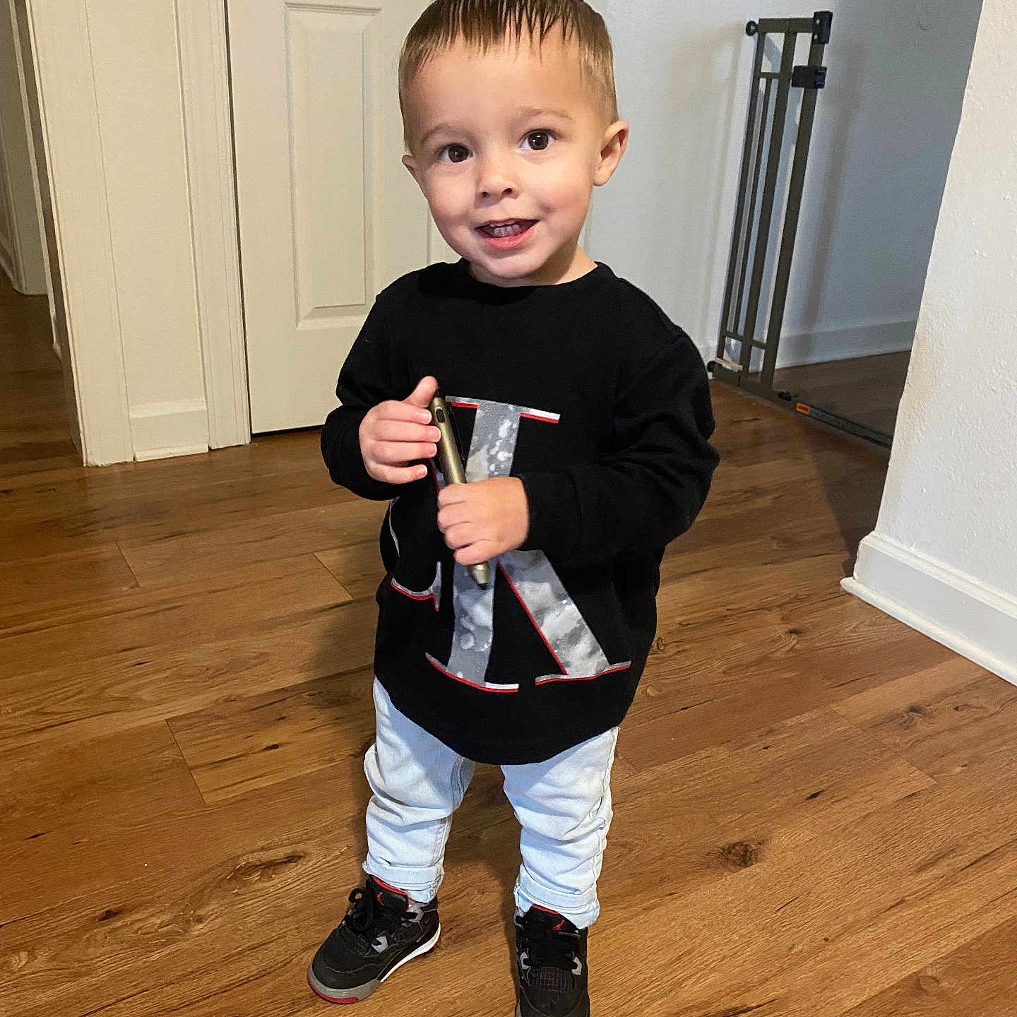 Tunney is registered to the contest to win money with this photo: baby_gate, black_sweatshirt, casual, child, clothing, door, flooring, happy, home, indoor, light_jeans, person, phone, portrait, smiling, sneakers, standing, toddler, wall, wooden_floor