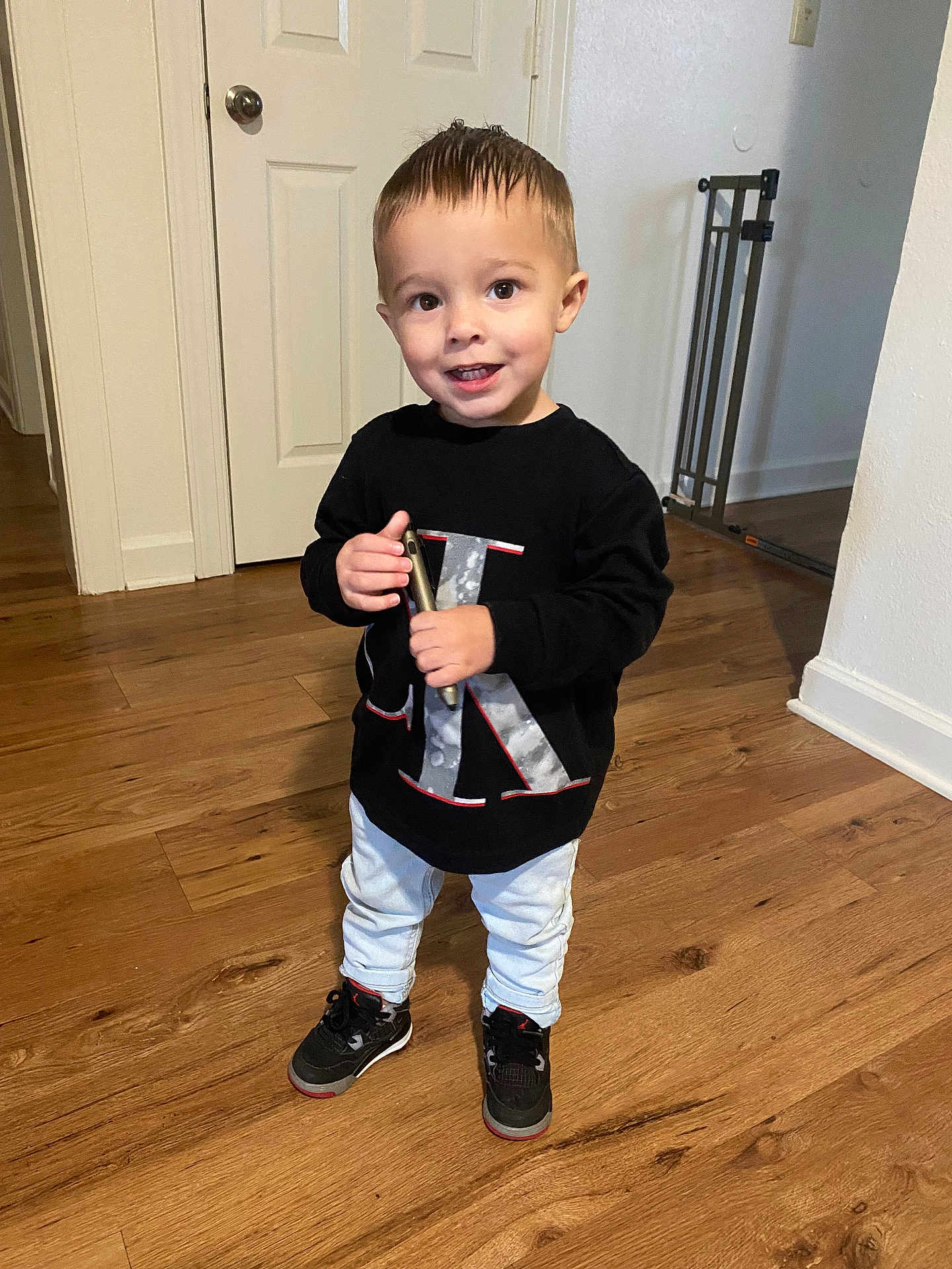 Tunney is registered to the contest to win money with this photo: child, toddler, indoor, wooden_floor, black_sweatshirt, light_jeans, sneakers, phone, smiling, standing, door, baby_gate, wall, flooring, clothing, person, portrait, happy, casual, home
