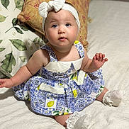 Mali is registered to the contest to win money with this photo: baby, child, blue_eyes, headband, dress, patterned_clothing, sitting, bed, pillow, white_socks, indoor, curious, cute, infant, soft_lighting, portrait, human, young_child, fabric, home