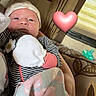 Adaline is registered to the contest to win money with this photo: baby, newborn, blanket, clothing, hat, mittens, pacifier, couch, curtains, heart_emoji, striped_clothing, person, infant, indoor, soft_toys, patterned_fabric, face, cozy, resting, home