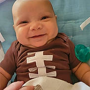 Jackson is registered to the contest to win money with this photo: baby, smiling, infant, brown_shirt, blue_cushion, pacifier, hand, blanket, cute, happy, face, portrait, child, indoor, closeup, person, seat, comfort, skin, expression