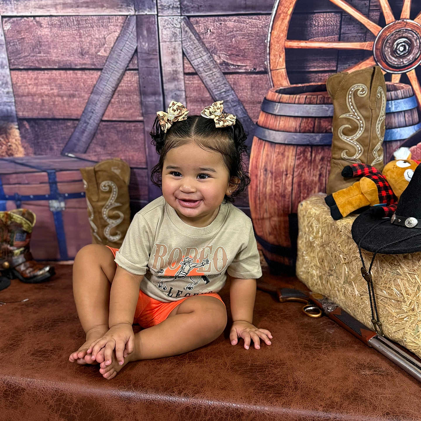 Myleiyah joined the competition — help win amazing prizes! barrel, bows, child, cowboy_boots, cowboy_hat, curly_hair, cute, happy, hay_bale, indoor, leather_surface, orange_shorts, playful, portrait, sitting, smiling, toddler, toy_rifle, tshirt, wooden_background