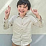 child, boy, smiling, peace_sign, hands, long_sleeve_shirt, buttoned_shirt, brown_pants, standing, wall, textured_wall, natural_light, shadow, happy, casual_clothing, portrait, outdoor, person, cute, youth