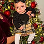 Maddie joined the competition — help win amazing prizes! baby, child, female, black_bow, black_top, plaid_skirt, white_tights, black_shoes, christmas_tree, ornaments, holiday, festive, decorations, lights, person, holding, indoor, celebration, smile, cute