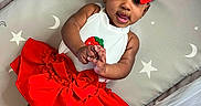 Island is registered to the contest to win money with this photo: baby, child, red_bow, red_skirt, white_top, playpen, stars, moons, smiling, cute, infant, clothing, hands, face, headwear, indoors, portrait, lying_down, curly_hair, skin