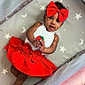 baby, child, clothing, curly_hair, cute, face, hands, headwear, indoors, infant, lying_down, moons, playpen, portrait, red_bow, red_skirt, skin, smiling, stars, white_top