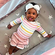 Island is registered to the contest to win money with this photo: baby, child, playpen, headband, bow, striped_shirt, pink_shorts, socks, smiling, tongue_out, lying_down, star_pattern, moon_pattern, cute, infant, happy, indoor, colorful_clothing, soft_toys, cozy