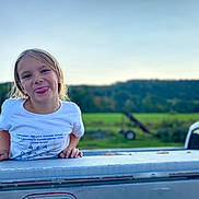 Katelyn is registered to the contest to win money with this photo: child, playful, tongue_out, white_tshirt, outdoor, green_field, sky, nature, daylight, smiling, person, young, casual_clothing, blonde_hair, fun, cheeky, metal_surface, background, vehicle, landscape
