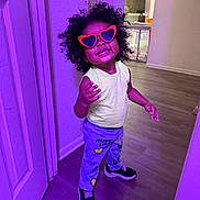 Maliyah is registered to the contest to win money with this photo: child, curly_hair, cute, door, fashion, happy, heart_shaped_glasses, home_interior, indoor, person, playful_pose, printed_pants, purple_light, smiling, sneakers, sunglasses, toddler, wall, wood_floor, yellow_tank_top