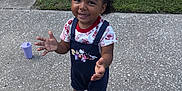 Maliyah is registered to the contest to win money with this photo: casual_clothing, child, concrete, cup, curly_hair, daylight, front_yard, grass, happy, outdoor, overalls, person, playful, portrait, shoes, shorts, smiling, socks, standing, toddler