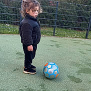 Lyam a rejoint le concours — aidez-le/la à gagner de superbes lots ! child, toddler, curly_hair, black_jacket, black_pants, sneakers, soccer_ball, outdoor, sports_field, green_surface, fence, grass, trees, playing, standing, focused, side_view, young_child, casual_clothing, daytime