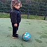 child, toddler, curly_hair, black_jacket, black_pants, sneakers, soccer_ball, outdoor, sports_field, green_surface, fence, grass, trees, playing, standing, focused, side_view, young_child, casual_clothing, daytime