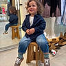 child, toddler, curly_hair, jeans, sneakers, jacket, stool, mirror, reflection, clothing_store, boots, hanging_clothes, floor, smiling, indoor, casual_wear, wooden_furniture, person, shopping, happy