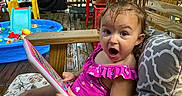 Taylor is registered to the contest to win money with this photo: child, toddler, girl, portrait, surprised, open_mouth, sitting, deck, patio, outdoor, sunlight, garden, plants, kiddie_pool, pool_toys, deck_chair, cushion, pink_dress, drawing_board, backyard