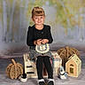 child, portrait, smiling, sitting, autumn, studio, wicker_pumpkin, handbag, plush_toy, miniature_house, bench, black_clothing, skirt, white_shoes, striped_socks, brown_hair, bangs, indoor, decor, woodland_theme