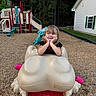 child, girl, toddler, playground, playground_equipment, frog_ride, outdoor, smiling, happy, portrait, sitting, hands_on_cheeks, blonde_hair, long_hair, black_shirt, mulch, forest, house, slide, recreation