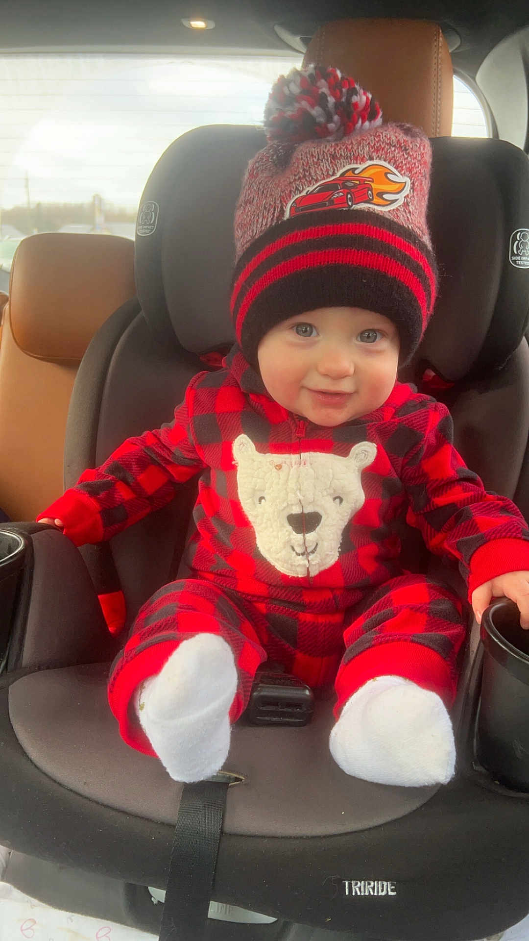 Noah is registered to the contest to win money with this photo: baby, child, car_seat, hat, pom_pom, onesie, red_and_black, plaid, smiling, socks, car_interior, seatbelt, face, winter_clothing, cute, infant, vehicle, window, daylight, portrait