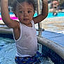 child, toddler, water, pool, swimwear, tank_top, curly_hair, splashing, outdoor, summer, sunlight, water_reflection, playful, swimming_pool, colorful_clothing, young_child, bare_arms, wet_clothes, recreation, fun