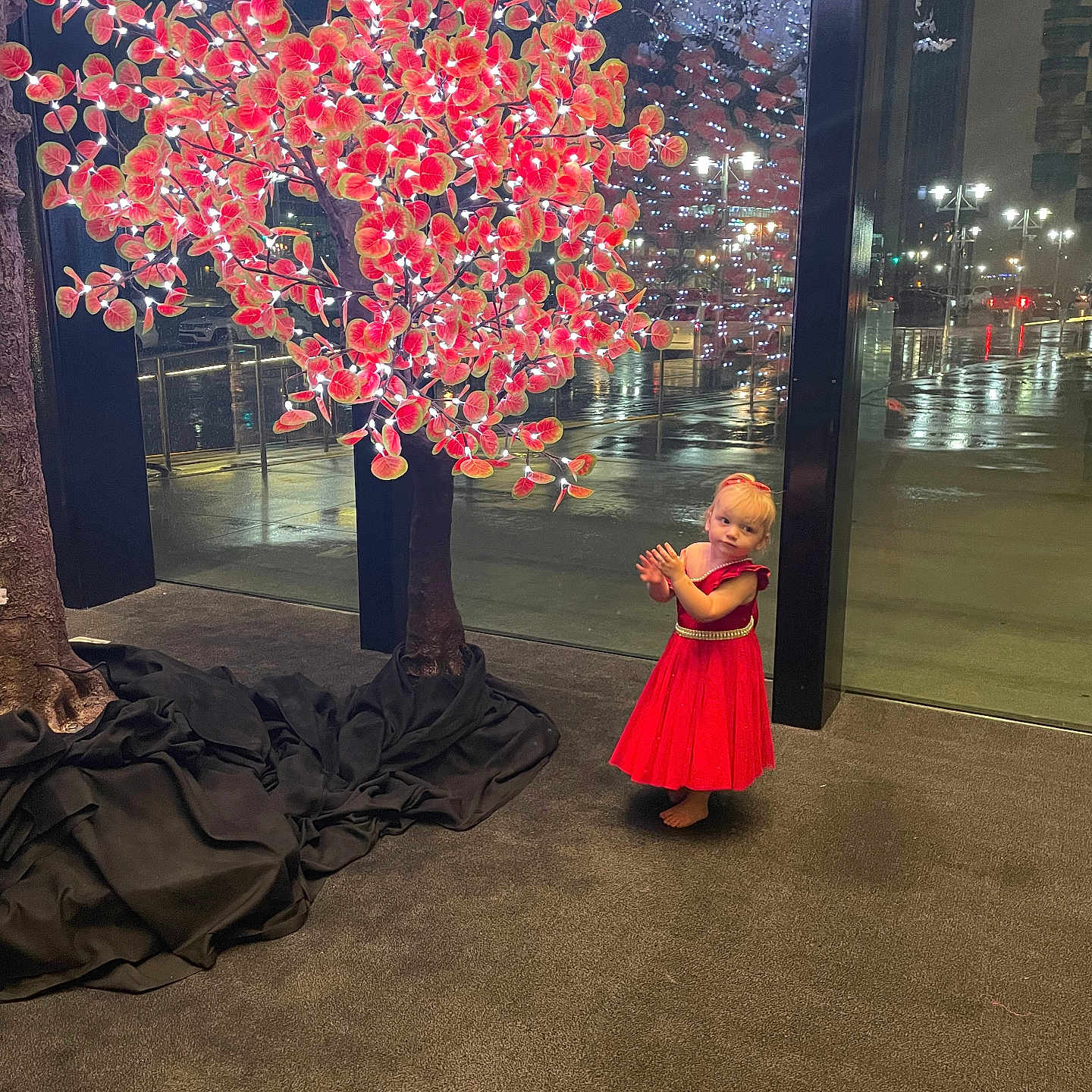 Regina joined the competition — help win amazing prizes! artificial_tree, barefoot, carpet, child, clapping, curious, decorative_tree, floor, glass_wall, illuminated, indoor, lights, night, rainy, red_dress, reflection, toddler, urban, window, young_girl