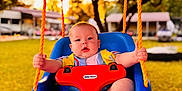 Eli is registered to the contest to win money with this photo: baby, child, swing, playground, outdoor, sunset, orange_rope, blue_swing, red_safety_bar, grass, trees, daylight, infant, person, seat, fun, cute, nature, summer, play