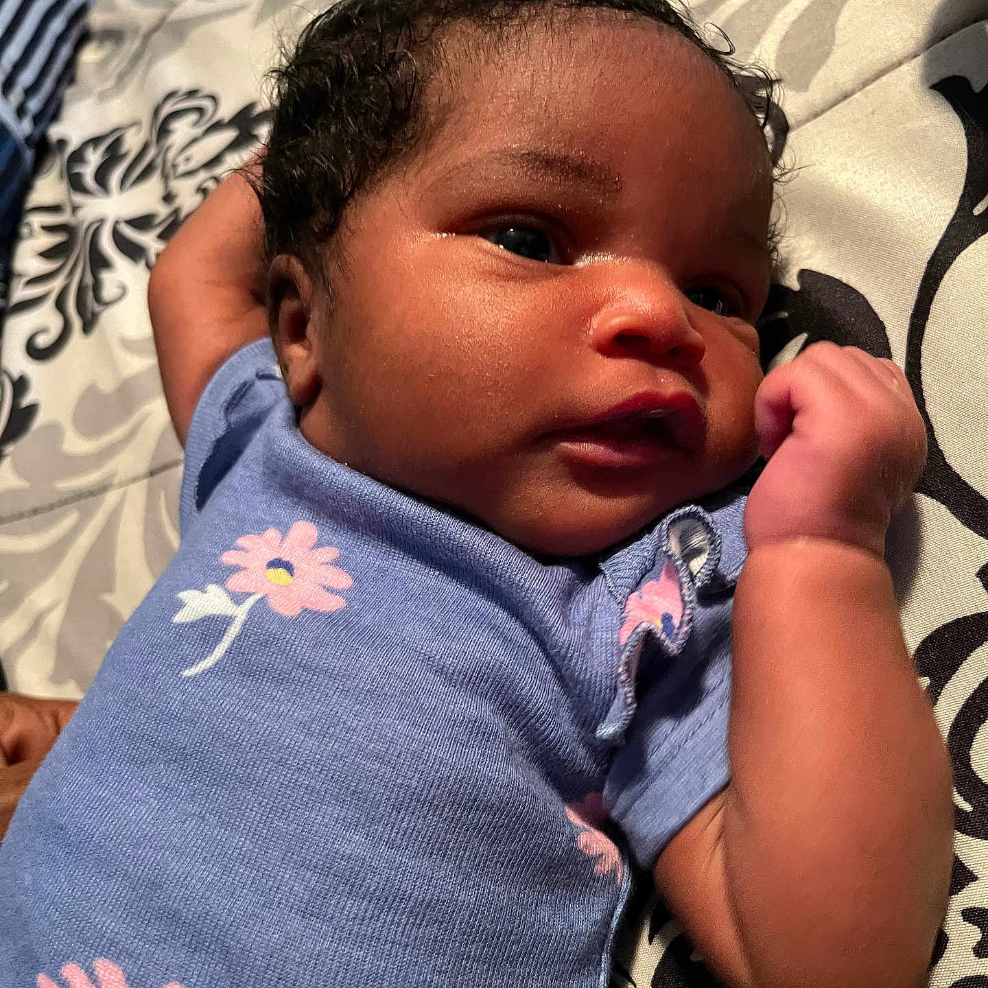 Honestly joined the competition — help win amazing prizes! baby, blue_onesie, child, closeup, curly_hair, cute, expression, face, fist, flowers, indoors, infant, lying_down, newborn, patterned_background, person, portrait, skin, small_hand, soft_light