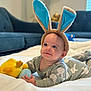 baby, bunny_ears, plush_toy, blanket, laying_down, indoors, cute, child, soft_toys, smiling, face, person, living_room, couch, cozy, headwear, infant, happy, clothing, portrait