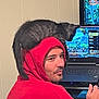 animal, black_cat, casual, cat_on_shoulder, companion, computer, face, game, gaming, hood, indoor, laptop, leisure, man, person, pet, red_hoodie, relaxation, screen, technology
