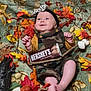 baby, child, smiling, camo_overalls, headband, autumn_leaves, pumpkin, chocolate_bar, blanket, fall_theme, decorations, marshmallows, stick, mushroom_pattern, orange, yellow, red, cute, happy, indoors