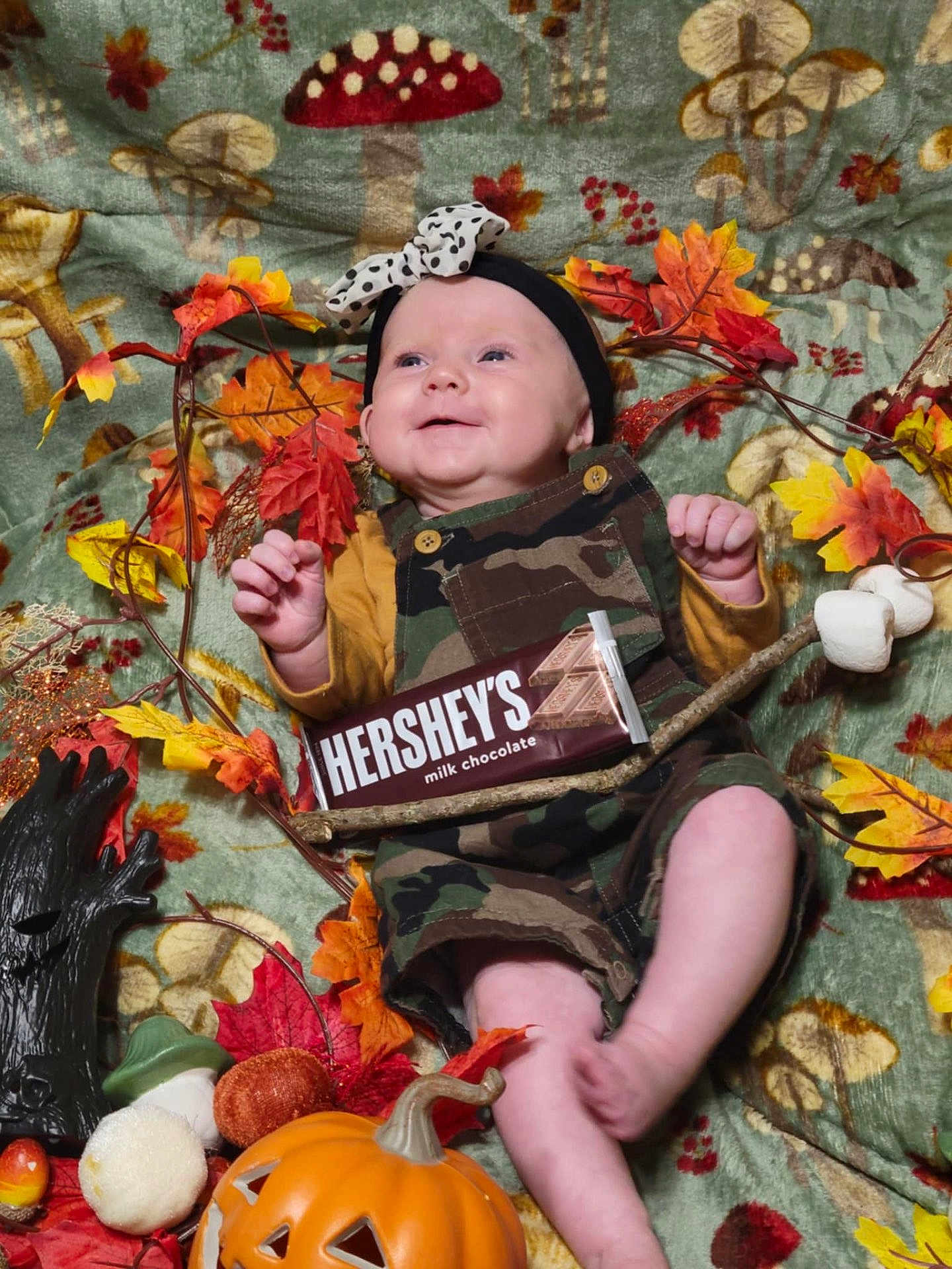 Berkley is registered to the contest to win money with this photo: baby, child, smiling, camo_overalls, headband, autumn_leaves, pumpkin, chocolate_bar, blanket, fall_theme, decorations, marshmallows, stick, mushroom_pattern, orange, yellow, red, cute, happy, indoors