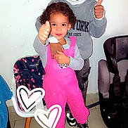 Elyäna a rejoint le concours — aidez-le/la à gagner de superbes lots ! child, kids, thumbs_up, smiling, indoor, overalls, pink_clothing, grey_sweater, chair, car_seat, floor_tiles, happy, portrait, standing, curly_hair, sneakers, positive_gesture, family, siblings, home