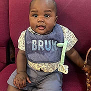 Kayden is registered to the contest to win money with this photo: baby, child, bib, pacifier, chair, red_chair, clothing, shorts, patterned_shirt, face, hand, cute, indoor, seated, expression, curious, skin, furniture, accessory, portrait