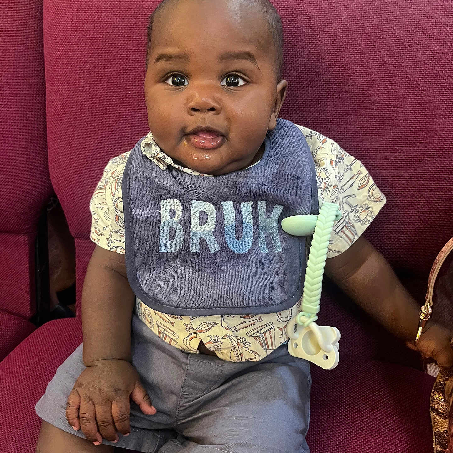 Kayden is registered to the contest to win money with this photo: baby, child, bib, pacifier, chair, red_chair, clothing, shorts, patterned_shirt, face, hand, cute, indoor, seated, expression, curious, skin, furniture, accessory, portrait