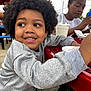 child, curly_hair, smiling, outdoor, picnic_table, red_table, grey_sweater, ice_cream, happy, sitting, food, casual, daylight, person, young, snack, fun, afro, candid, background