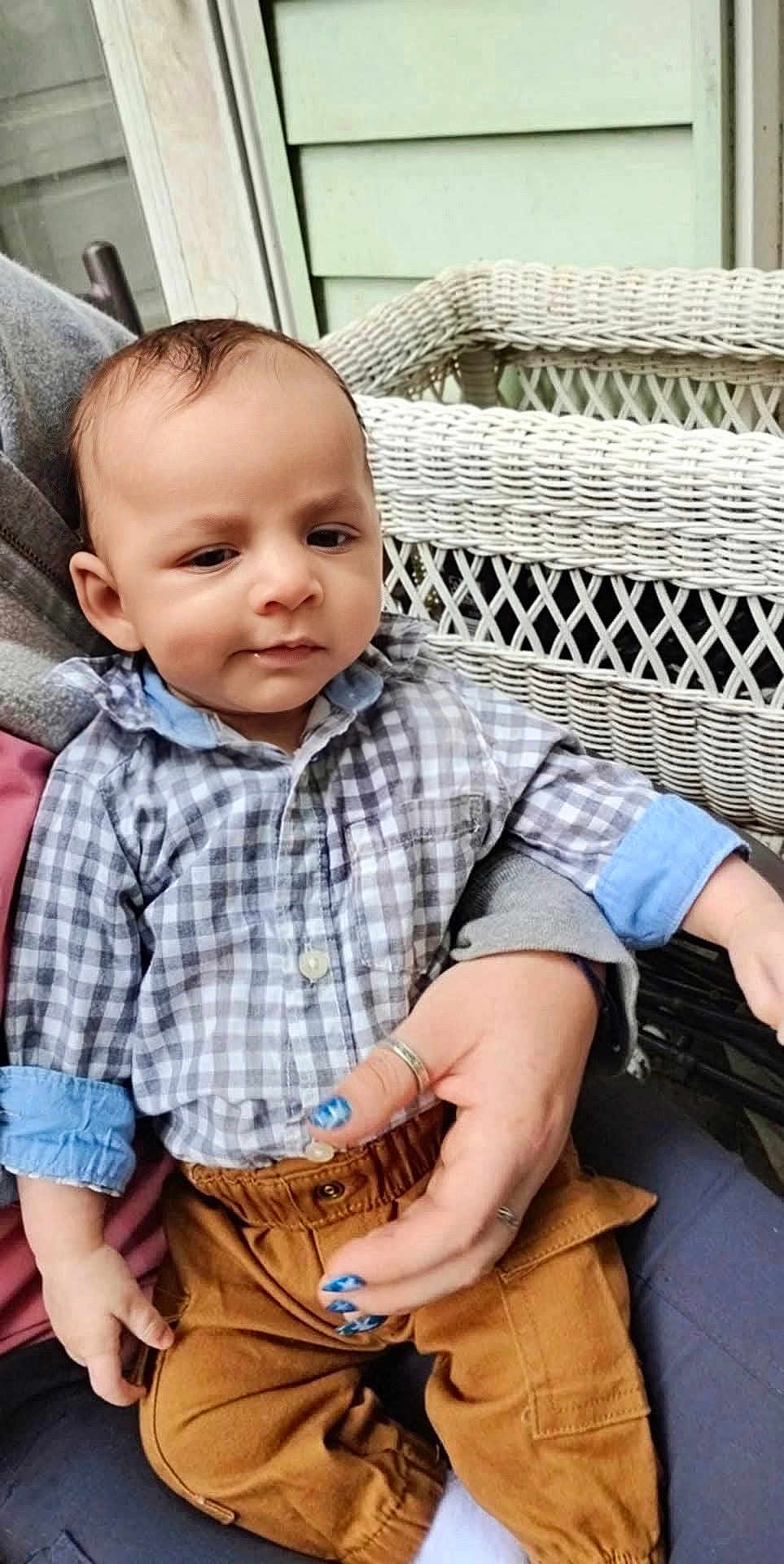 Izayah is registered to the contest to win money with this photo: baby, child, person, checkered_shirt, brown_pants, hand, nail_polish, wicker_chair, outdoor, green_siding, sitting, casual_clothing, portrait, infant, cute, holding, expression, fingernails, fabric, relaxed