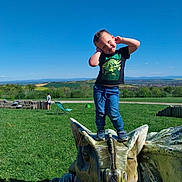 Louka participe au concours pour gagner de l'argent avec cette photo : child, boy, dinosaur_sculpture, outdoor, park, grass, blue_sky, smiling, playful, casual_clothing, sculpture, nature, sunny_day, greenery, toy, landscape, recreation, posing, happy, scenic_view