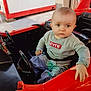 baby, child, toy_car, red, sweater, jeans, seatbelt, indoor, person, hand, face, steering_wheel, casual, curious, seated, clothing, background, man, phone, expression