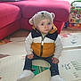toddler, child, hat, knit_hat, vest, puffer_vest, jeans, white_shoes, indoor, play_mat, toy, walker, red_couch, pillow, floor, curtains, cute, sitting, portrait, young_child
