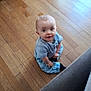 baby, child, floor, wooden_floor, blue_clothing, pants, shirt, sitting, indoor, curious, looking_up, couch, furniture, home, smile, person, infant, young_child, casual, portrait