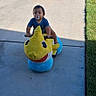child, toddler, baby, dinosaur_toy, ride_on_toy, pacifier, outdoor, driveway, sidewalk, grass, sunlight, shadow, striped_shirt, seated, portrait, playtime, toy, yellow, blue, residential
