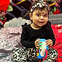 Harini is registered to the contest to win money with this photo: toddler, child, toy, phone, sitting, leopard_print, headband, black_clothing, smiling, indoor, bedspread, colorful_background, patterned_pants, grey_socks, cute, portrait, happy, baby, earrings, playful