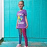 child, standing, purple_shirt, pokemon, trainer, leggings, leopard_print, colorful_shoes, high_tops, outdoor, concrete_floor, blue_wall, casual_clothing, smiling, young_girl, fashion, portrait, vibrant_colors, playful, confident
