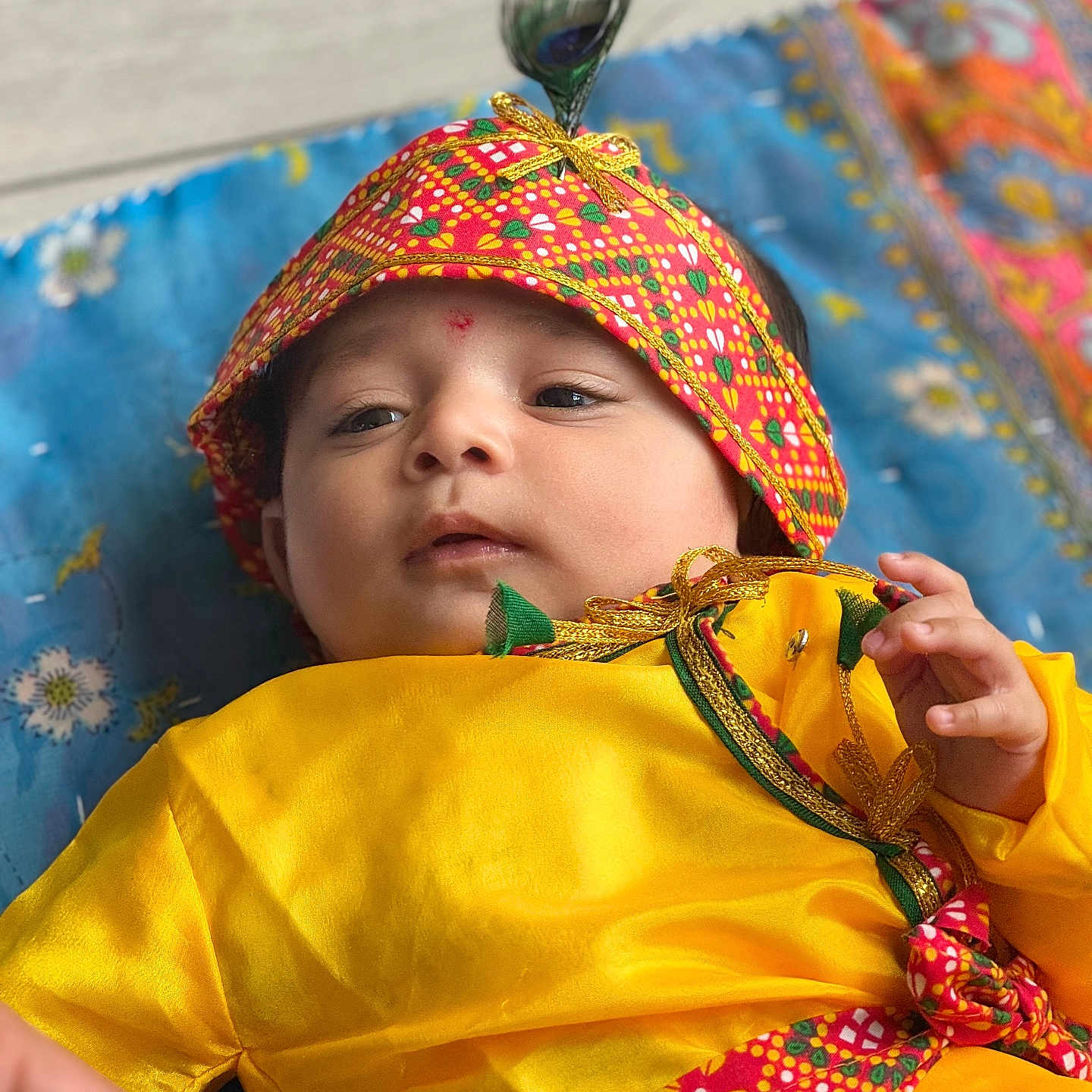 Parth is registered to the contest to win money with this photo: baby, blue_blanket, child, colorful, cute, expression, face, feather, floral_pattern, hand, headband, indoors, infant, lying_down, patterned_fabric, person, portrait, soft_light, traditional_clothing, yellow_clothing