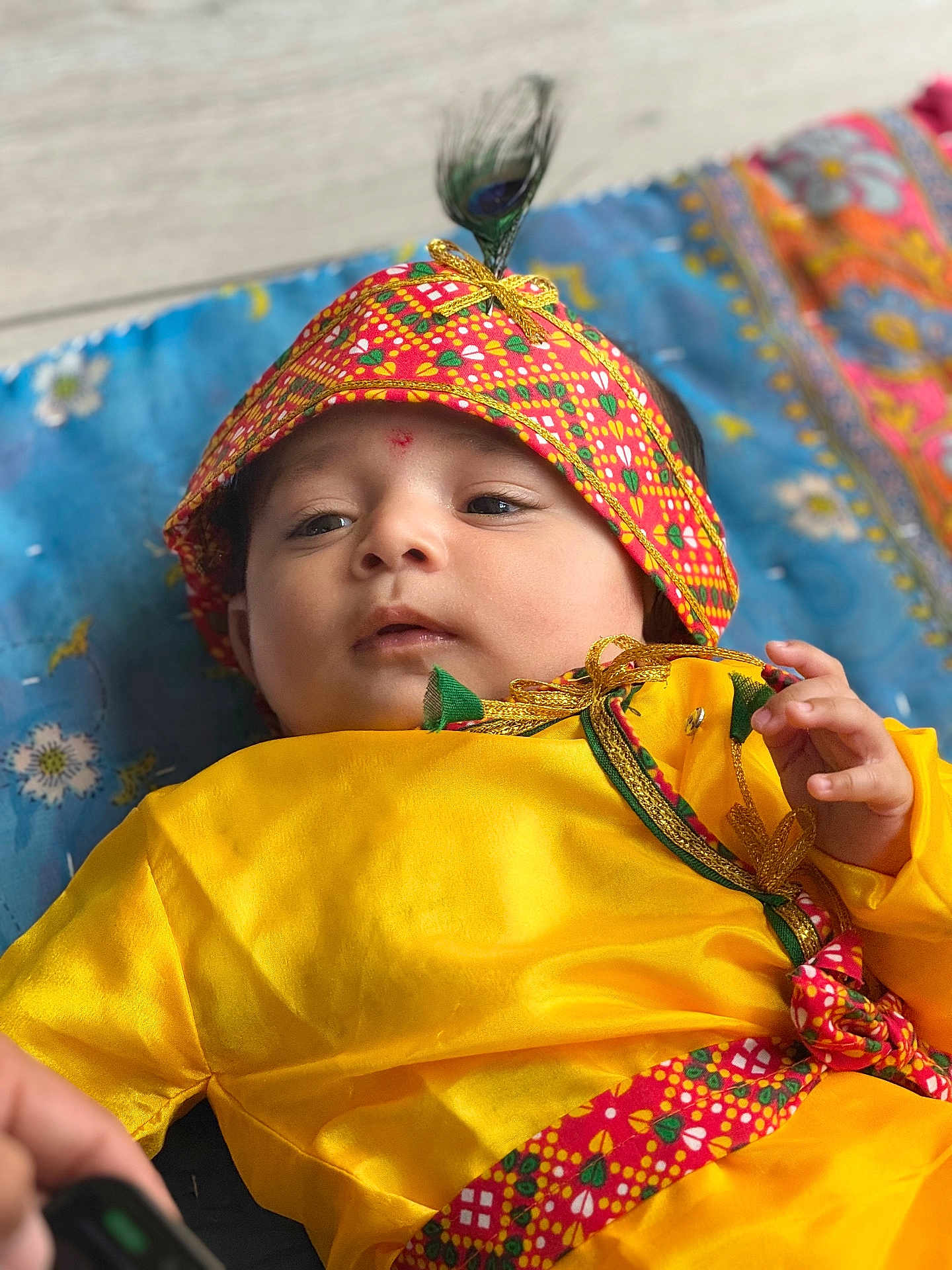 Parth is registered to the contest to win money with this photo: baby, child, yellow_clothing, headband, feather, patterned_fabric, blue_blanket, floral_pattern, traditional_clothing, infant, portrait, cute, lying_down, hand, face, expression, indoors, colorful, soft_light, person