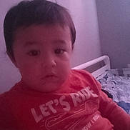 Kayden joined the competition — help win amazing prizes! child, toddler, face, hair, eyes, red_shirt, text_on_shirt, bed, bedroom, blanket, pillow, wall, indoor, sitting, portrait, neutral_expression, young_child, person, casual_clothing, cozy