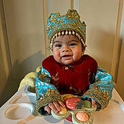 Iraj joined the competition — help win amazing prizes! baby, child, high_chair, toy, crown, blue_clothing, gold_embroidery, smiling, indoor, cute, portrait, hands, face, person, young_child, playing, happy, seated, colorful_toy, traditional_clothing