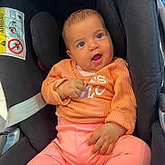 Nayla participe au concours pour gagner de l'argent avec cette photo : baby, child, infant, car_seat, orange_sweater, pink_pants, white_socks, smiling, happy, indoors, seatbelt, safety_label, person, cute, portrait, young_child, clothing, head, hand, face